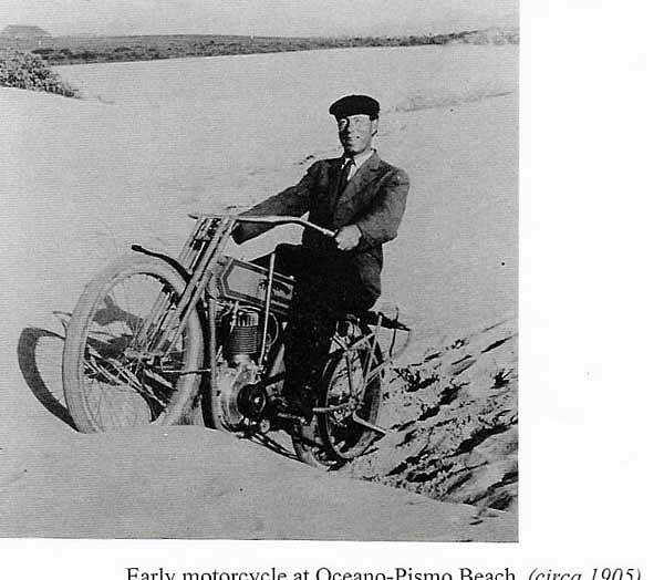 A black and white photo of a man on an old harley davidson motorcycle on Pismo beach dated 1905