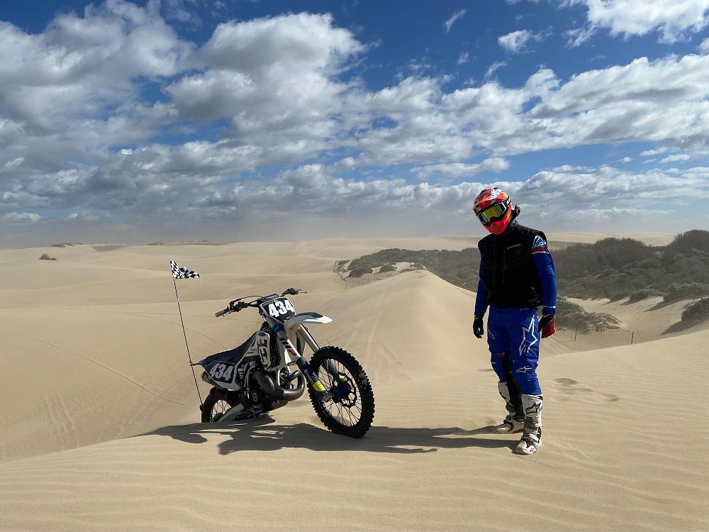 A dirt bike rider has his bike parked in the deep dune sand with a dust cloud in background