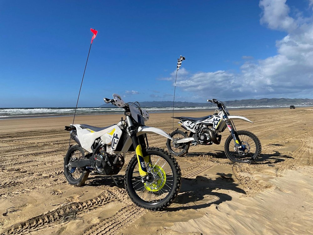 Two dirtbikes parked on the shoreline, with the waves and blue sky in the background