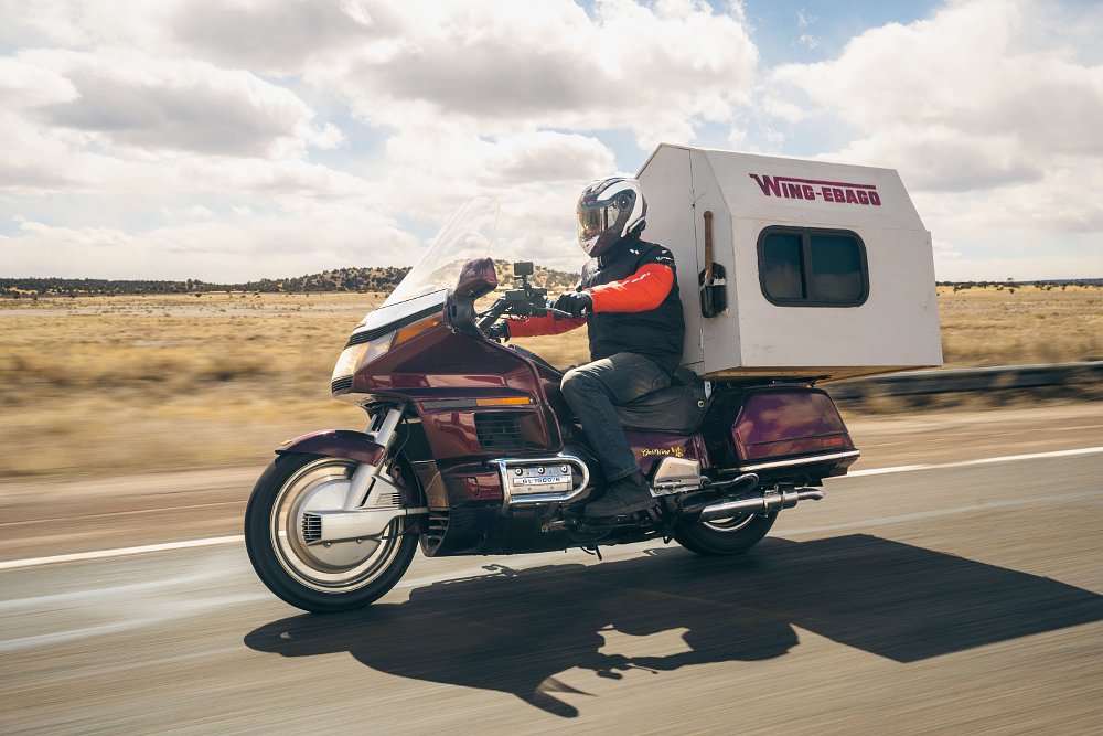 A 1989 Honda Gold Wing with a tiny house on the back, being ridden down a paved road in the desert.