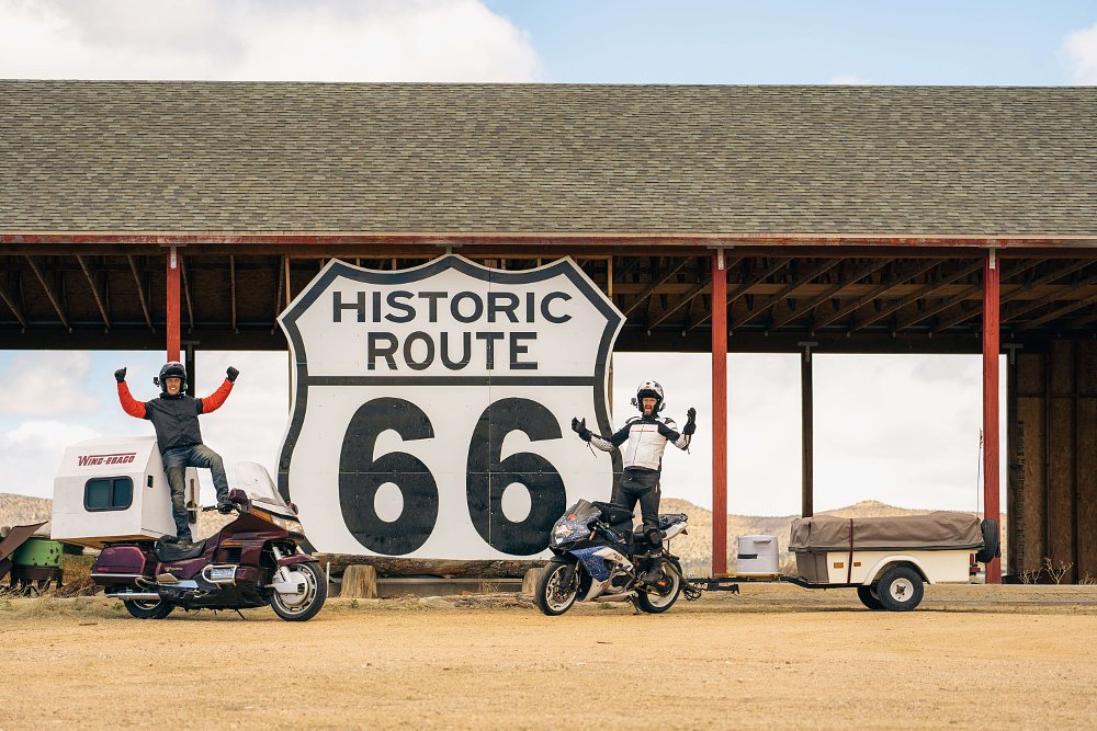 Two men pose with their motorcycle motorhomes next to a giant Route 66 sign.