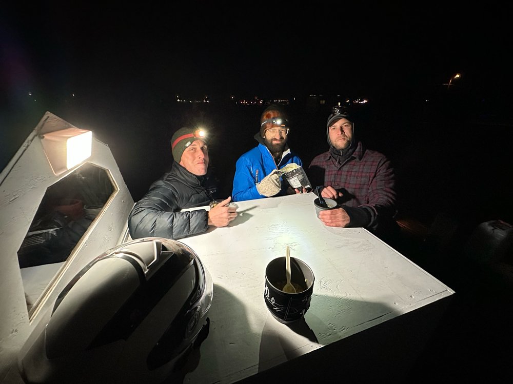 Three people eating freeze-dried food in a campsite at night.