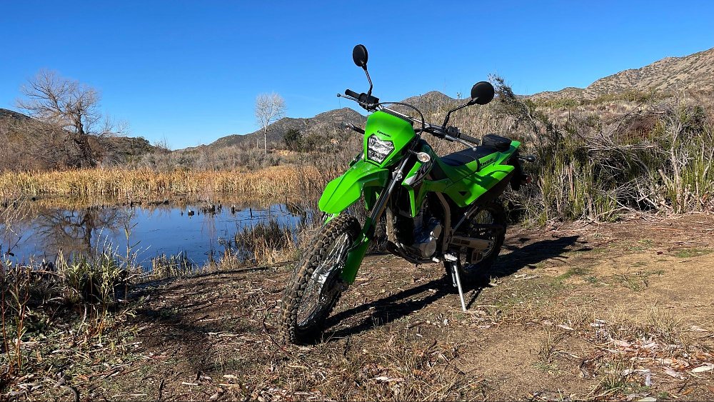 A bright green KLX300 parked in front of a blue duck pond with ducks swimming around