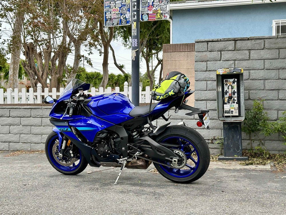 A 2024 Yamaha R1 with a bundle of apparel strapped to the back seat, parked in front of a pay phone.