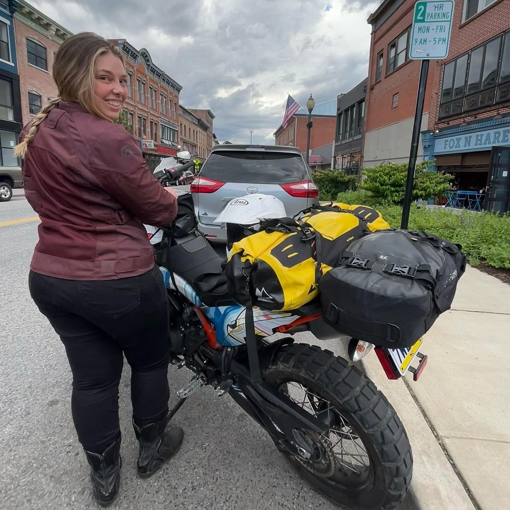 Liz beside her KTM motorcycle loaded with gear on a small town street