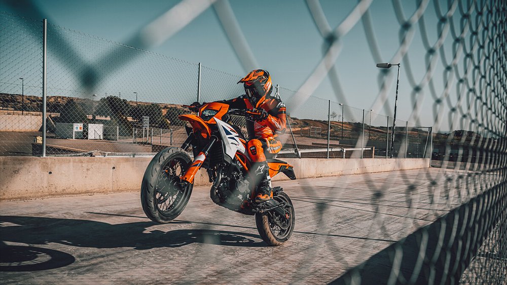 A rider pops a wheelie aboard the KTM 390 SMC R with a chainlink fence in the foreground.
