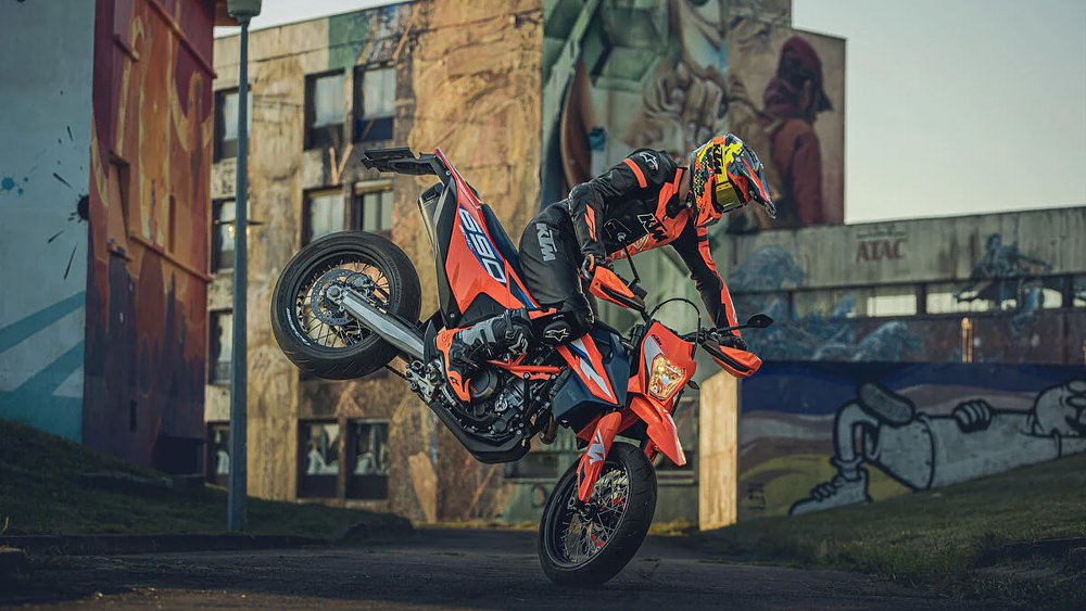 A KTM 690 SMC R rider performs a nose wheelie on an urban street.