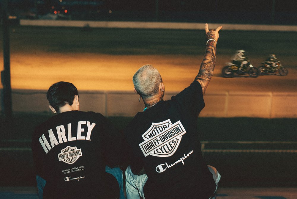 two fans wearing Harley-Davidson shirts watching a flat-track race at night
